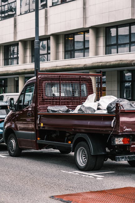 A burgundy flatbed van parked on a city street in Whitechapel, with a large open cargo area loaded with various household items including cardboard boxes, wrapped furniture, and plastic bags. The cargo is secured with straps, and the van features a metal grid behind the driving cab to protect the load. The background shows modern office or residential buildings with large windows and curved architectural elements. The scene depicts the loading or transportation process typical of home relocation services, with the van positioned close to the pavement for easy access by [COMPANY_NAME] for furniture transport and moving logistics in Whitechapel.
