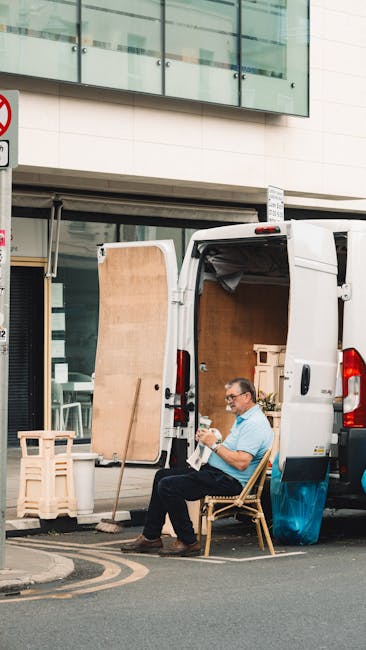 A man wearing glasses and a light blue shirt is seated on a wooden chair next to a white moving van parked outside a modern building with large glass windows. The van's rear doors are open, revealing an interior filled with cardboard boxes, wooden furniture panels, and packing materials. A large wooden panel leans against the van's open door, while a small wooden crate is positioned nearby. The man appears to be taking a break during a home relocation, with the outdoor pavement visible beneath him and some street signage partially seen in the background. The scene reflects the loading or unloading process typical of professional removal services, such as those offered by Man with Van Whitechapel, focusing on furniture transport and packing during a residential move.