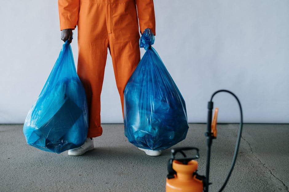 A person, dressed in orange work overalls and white shoes, is standing indoors on a concrete floor, holding two large blue plastic bags filled with bulky waste or recyclable materials. The individual's hands are gripping the bags, which are slightly translucent, revealing contents inside. The background features a plain light grey wall, with minimal additional objects visible. In the foreground, there is an orange and black handheld sprayer or similar equipment, connected via a hose to a container. The scene suggests a waste collection or preparation process associated with home or office relocation services, such as packing and loading bulky waste for removal, which aligns with the services offered by Man with Van Whitechapel in house removals and moving logistics.