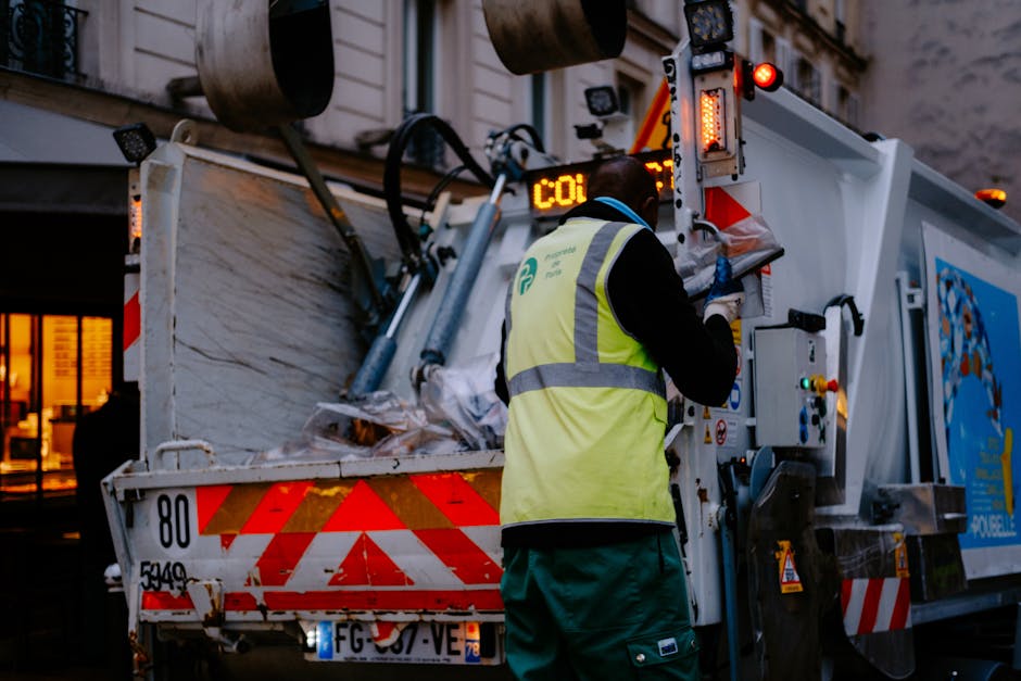 A worker wearing a high-visibility vest and dark clothing operates a waste collection vehicle during a home relocation or clearance process. The vehicle is parked outside a building with a partially visible brick wall and a window illuminated with warm light. Inside the open rear compartment of the waste collection truck, various items including crumpled cardboard boxes, plastic-wrapped materials, and debris are visible, indicating the collection of bulky waste. The worker is standing on pavement, facing the open rear of the truck, and is using controls to manage the loading process. The scene is set during low light conditions, possibly early evening, with artificial lighting highlighting the vehicle's equipment and the worker's actions. This scenario exemplifies waste or rubbish removal in the context of house removals or property clearance, supported by [manwithvanwhitechapel.co.uk], which offers professional removals and clearance services in Whitechapel E1.