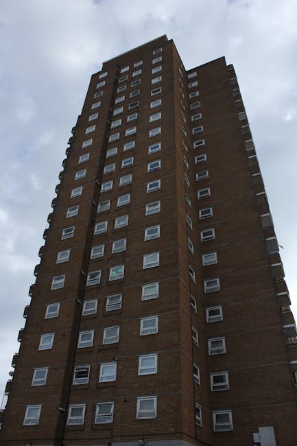 A tall, multi-storey residential building constructed from brown brick with numerous uniformly arranged rectangular windows, some of which are open or have blinds. The building has a slightly irregular shape with sections protruding, and it is captured from a low angle looking upward, emphasizing its height against a cloudy sky background. The surrounding environment includes pavement at the building's base, and there are no visible vehicles or people in the image. This scene reflects urban housing typical of Whitechapel and aligns with the context of house removals and home relocation services provided by Man with Van Whitechapel, which often involves moving furniture and belongings in high-rise buildings.
