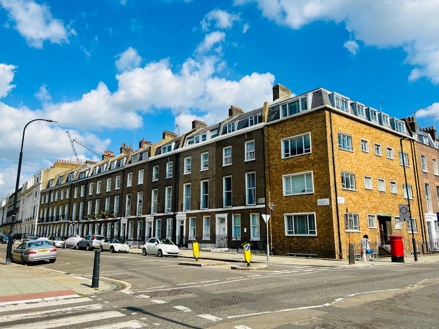 A four-storey brick residential building situated on a street corner in Whitechapel, London, under a partly cloudy blue sky. The building has multiple rows of white-framed windows, with some featuring small balconies or window boxes. The ground floor includes front doors with small steps leading to the pavement. Several parked cars are along the street, accompanied by bollards and roadside signs. A person is walking on the pavement near the building entrance, and a red post box is positioned at the corner. The scene captures typical urban residential architecture, with clear weather providing natural lighting suitable for a home relocation or furniture transport operation, as handled by Man with Van Whitechapel's removals team.