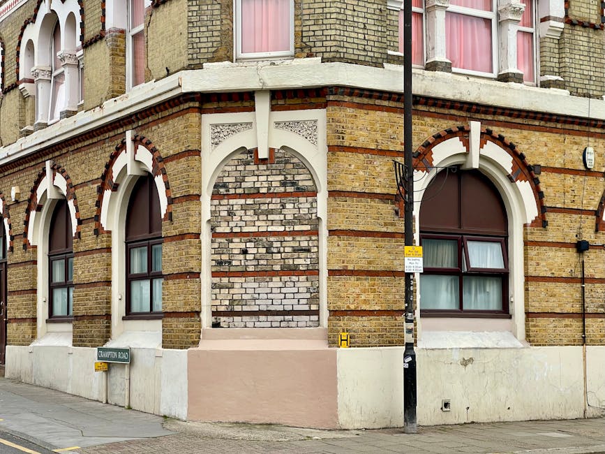 A tall, multi-storey residential building constructed from brown brick with numerous uniformly arranged rectangular windows, some of which are open or have blinds. The building has a slightly irregular shape with sections protruding, and it is captured from a low angle looking upward, emphasizing its height against a cloudy sky background. The surrounding environment includes pavement at the building's base, and there are no visible vehicles or people in the image. This scene reflects urban housing typical of Whitechapel and aligns with the context of house removals and home relocation services provided by Man with Van Whitechapel, which often involves moving furniture and belongings in high-rise buildings.