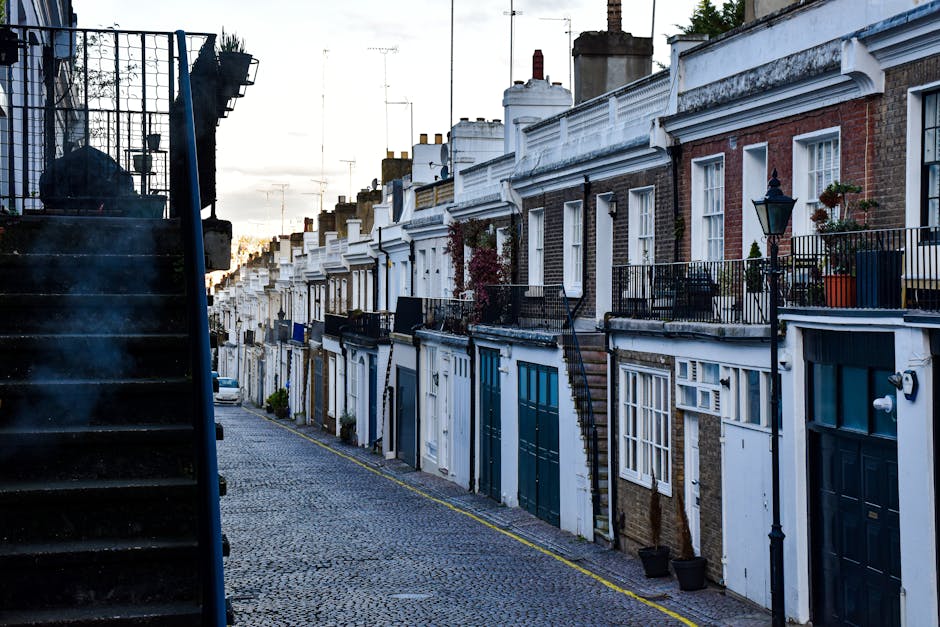 An exterior view of a narrow cobblestone street in Whitechapel lined with traditional terraced houses featuring brick facades, white wooden window frames, decorative cornices, and small private balconies with black wrought iron railings. Several potted plants and flower boxes adorn the balconies, and a classic street lamp is attached to one of the properties. The photograph is taken during daylight with a partly cloudy sky, capturing the urban setting typical of London’s Victorian residential area. The perspective shows the street extending towards the horizon, emphasizing its tight and historic character. This setting is representative of the type of property where Man with Van Whitechapel provides house removal services, including navigating narrow staircases and tight entrances during home relocation and furniture transport processes.