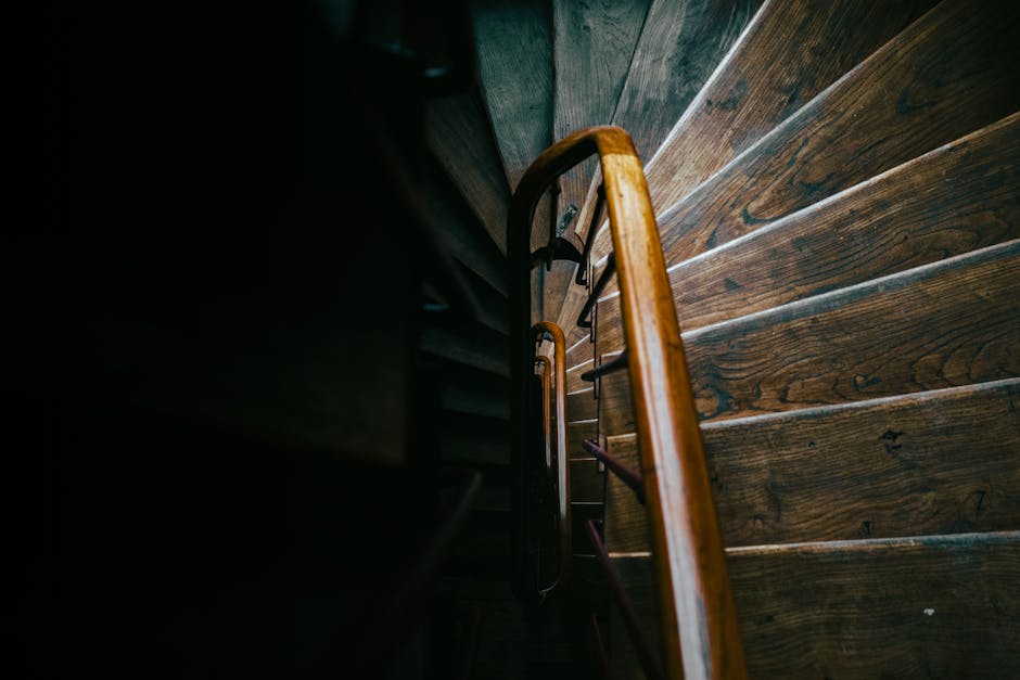 A view looking down a narrow wooden staircase inside a historic residential property, showing dark wooden steps and a polished wooden handrail. The staircase appears steep and tight, characteristic of Victorian-era homes. The photograph captures the top of the staircase from an overhead perspective, with the steps descending into shadow below. The environment is dimly lit, highlighting the natural wood grain and finish on the stairs and handrail. This setting illustrates the challenging nature of navigating narrow, steep Victorian stairs during a house removal or home relocation process. Occasionally, professionals from Man with Van Whitechapel may be called upon to handle the careful moving of furniture and boxes, including packing and transport through confined staircases like these. The image reflects the logistics involved in furniture transport and the importance of careful planning during the loading process for house removals.