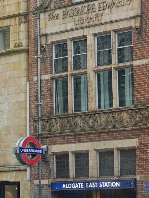 An exterior view of a narrow cobblestone street in Whitechapel lined with traditional terraced houses featuring brick facades, white wooden window frames, decorative cornices, and small private balconies with black wrought iron railings. Several potted plants and flower boxes adorn the balconies, and a classic street lamp is attached to one of the properties. The photograph is taken during daylight with a partly cloudy sky, capturing the urban setting typical of London’s Victorian residential area. The perspective shows the street extending towards the horizon, emphasizing its tight and historic character. This setting is representative of the type of property where Man with Van Whitechapel provides house removal services, including navigating narrow staircases and tight entrances during home relocation and furniture transport processes.