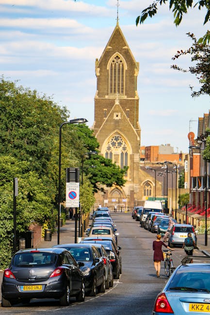 A narrow urban street lined with parked cars on both sides, with a woman walking and riding a bicycle along the pavement. Green trees border the street, and black lampposts are visible. In the background, a large historic stone church with a tall, pointed steeple, arched windows, and Gothic architectural features dominates the scene. The sky is partly cloudy with patches of blue. This setting depicts a quiet area suitable for house removals and furniture transport, where [COMPANY_NAME] may assist with home relocation or moving logistics during street and community access, with loading processes likely occurring in the vicinity of the church and nearby properties.