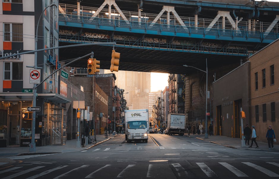 A city street intersection during daylight with a white delivery van positioned in the center of the road, facing toward the viewer, and several other trucks parked along the sides. Overhead, a large blue metal bridge spans the width of the street with supporting beams and signage visible. On either side of the street, there are multi-storey brick and glass buildings, some with commercial signage, and sidewalks with pedestrians walking past. Traffic lights hang above the intersection, and traffic signals are visible at the crossroads. The scene captures a typical urban environment suitable for a home relocation project, with the delivery van possibly being used for furniture transport or packing and moving services by Man with Van Whitechapel, in connection with house removals and logistical planning.