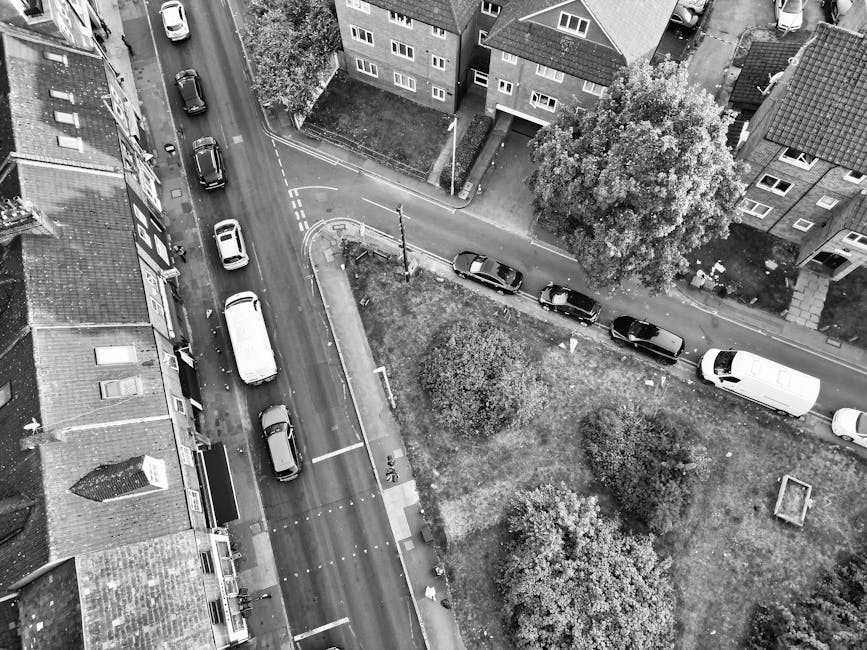 An aerial black-and-white photograph of a residential street scene showing several parked cars along a narrow road, adjacent to a small green park area with bushes and trees, and multi-story brick buildings with pitched roofs on either side. On the pavement, a man with a van from Man with Van Whitechapel is involved in home relocation activities, with cardboard boxes and wrapped furniture visible as part of the packing and moving process. The loading area near the white van features a trolley and moving blankets, demonstrating the logistics of furniture transport and loading during a house removal in Whitechapel, with the scene illuminated by natural daylight.