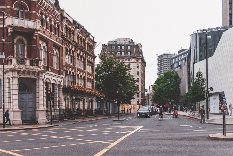 A city street intersection during daylight with a white delivery van positioned in the center of the road, facing toward the viewer, and several other trucks parked along the sides. Overhead, a large blue metal bridge spans the width of the street with supporting beams and signage visible. On either side of the street, there are multi-storey brick and glass buildings, some with commercial signage, and sidewalks with pedestrians walking past. Traffic lights hang above the intersection, and traffic signals are visible at the crossroads. The scene captures a typical urban environment suitable for a home relocation project, with the delivery van possibly being used for furniture transport or packing and moving services by Man with Van Whitechapel, in connection with house removals and logistical planning.