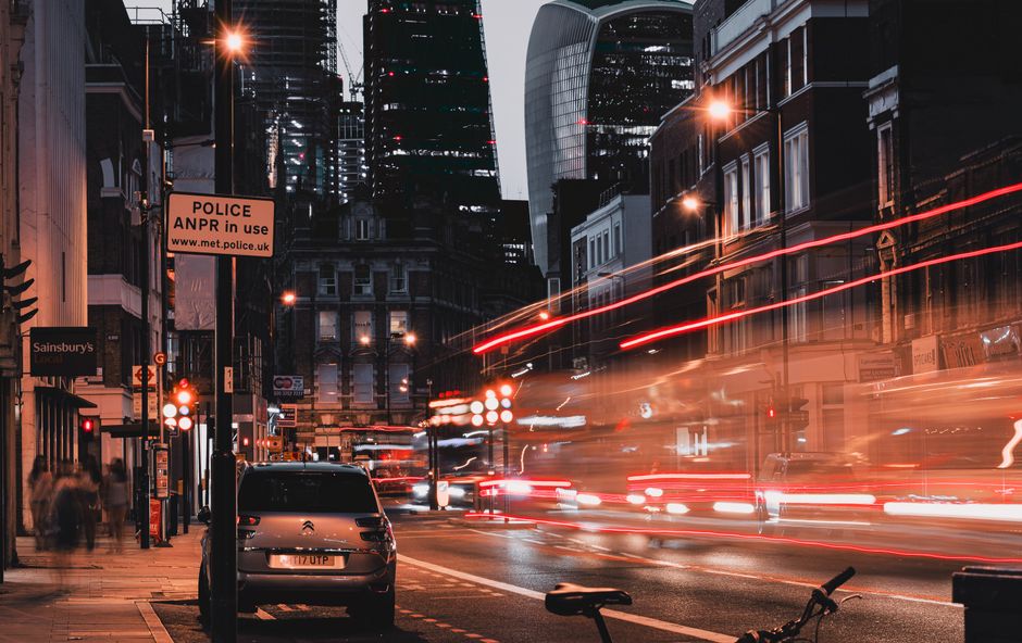 A nighttime urban street scene showing a city centre with tall modern skyscrapers and historic brick buildings along Commercial Road in Whitechapel. In the foreground, a parked car is situated near a pole displaying a parking suspension sign indicating police enforcement and parking restrictions relevant to house removals and logistics. The street is illuminated by streetlights and building lights, creating a warm glow. Light trails from passing vehicles suggest recent movement, with streaks of red and white light captured through long exposure. Pedestrians are visible on the sidewalk, and a van from Man with Van Whitechapel is partially visible, likely involved in a home relocation or furniture transport process nearby. The environment reflects an active, busy setting typical of a commercial district in London, supporting efficient packing and moving operations in coordination with parking suspensions for house removals.