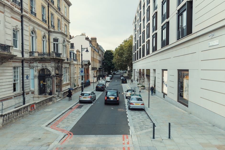 A wide street scene on Commercial Road, Whitechapel, showing parked cars on both sides of the road and a section of the pavement designated for loading and unloading with red and orange lines painted on the asphalt, indicating parking suspensions for moves. On the left, an ornate historic building with detailed stonework and a black metal gate is visible, while on the right, a modern building with large windows and a smooth facade extends along the sidewalk. Pedestrians are walking along the pavement, and a white van belonging to Man with Van Whitechapel is positioned further down the street, partially visible in the background. The environment is well-lit, with trees lining the street, and the scene captures the logistical context of home relocation activities, including furniture transport and packing processes involved in supported removals.