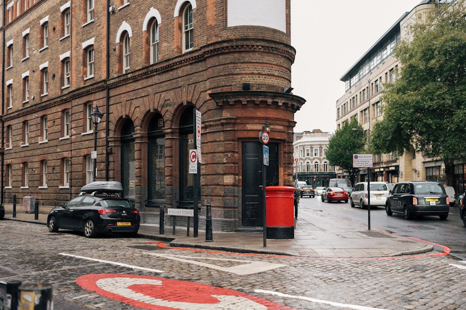 A nighttime urban street scene showing a city centre with tall modern skyscrapers and historic brick buildings along Commercial Road in Whitechapel. In the foreground, a parked car is situated near a pole displaying a parking suspension sign indicating police enforcement and parking restrictions relevant to house removals and logistics. The street is illuminated by streetlights and building lights, creating a warm glow. Light trails from passing vehicles suggest recent movement, with streaks of red and white light captured through long exposure. Pedestrians are visible on the sidewalk, and a van from Man with Van Whitechapel is partially visible, likely involved in a home relocation or furniture transport process nearby. The environment reflects an active, busy setting typical of a commercial district in London, supporting efficient packing and moving operations in coordination with parking suspensions for house removals.