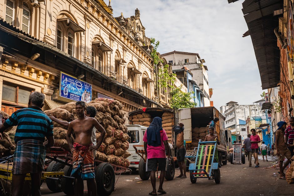 A busy urban street scene with several men and women engaged in loading and unloading large bags and boxes of goods onto and from small trucks with open backs, situated in a narrow, crowded street near Brick Lane Market in Whitechapel. The image shows a building with ornate architectural details and multiple windows on the left, with a billboard advertising beer fixed to the facade. The street is filled with activity, including people carrying items, pushing trolleys, and walking along the pavement, indicating an active home relocation or furniture transport process. The vehicles are parked close to the curb, and the area appears lively with a mix of residential and commercial buildings. This scene exemplifies typical moving logistics in an urban setting, which is a service provided by companies like Man with Van Whitechapel, specializing in removals and household moving services in the area.