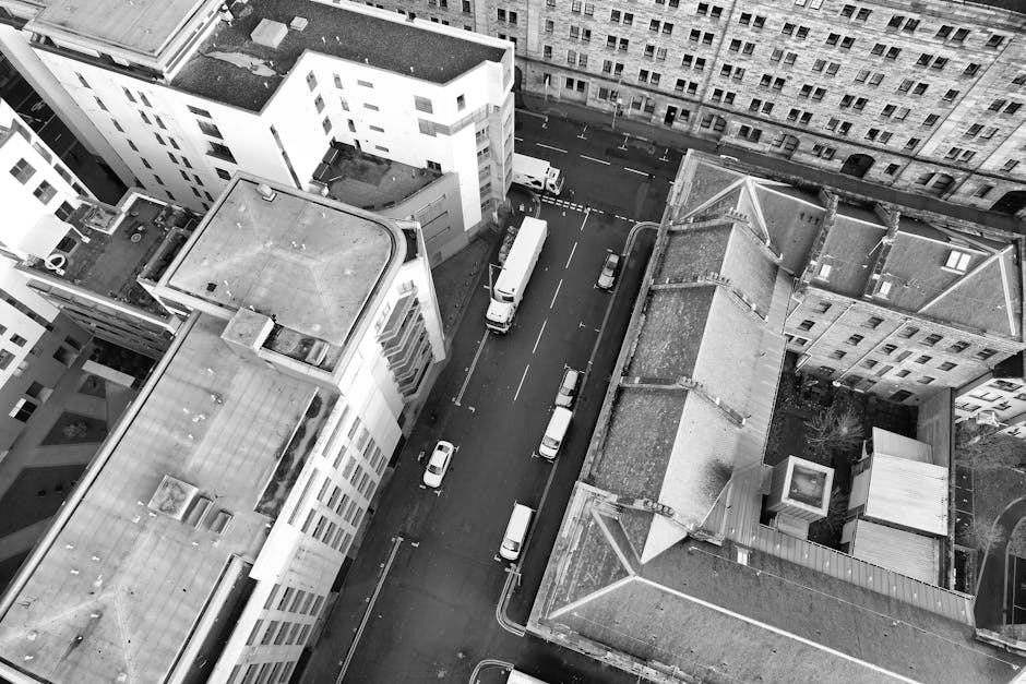 An aerial black-and-white photograph shows the rooftops and upper floors of several multi-storey buildings in Whitechapel, London, with a focus on a narrow street below. The street features parked cars along both sides, and a white moving truck is visible near the curb, partially loaded or unloaded with furniture and boxes. The scene captures the loading process involved in home relocation or furniture transport, with adjacent buildings constructed from brick and concrete, some with sloped or flat roofs. Man with Van Whitechapel's moving service may utilize such streets for efficient loading and unloading during same-day removals, as visible equipment like trolleys or blankets might be in use. The image emphasizes urban logistics, tight space maneuvering, and the equipment used for packing and moving within this residential area, highlighting the importance of careful planning in house removals in Whitechapel.