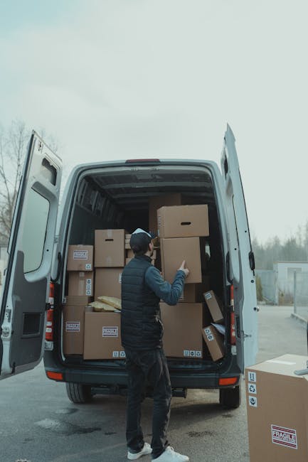 A man wearing a navy blue vest, black trousers, and a baseball cap is inside the back of a white moving van with its rear doors open, loading and arranging various brown cardboard boxes of different s