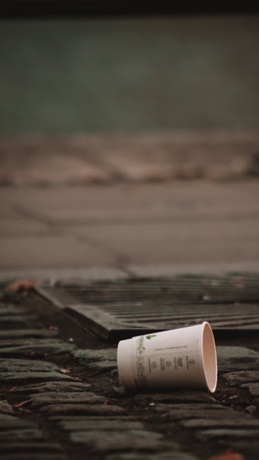 A white disposable paper cup is lying on its side on a weathered wooden doorstep outside a property, with the open end facing downward. The cup features printed text and branding, and is situated on a dark, textured doormat that is only partially visible. The surrounding area includes a black rubber threshold and a doorway with a dark frame, indicating the entrance to a home. In the background, a portion of the exterior wall with a muted green tint and horizontal lines is visible, suggesting siding or paneling. The lighting appears natural, capturing a quiet moment during the day. This scene relates to home relocation, as it depicts the process of packing or clearing items before or during a house move, with the casual placement of the cup hinting at packing and moving activities. The image is associated with professional removals and transport services such as those provided by Man with Van Whitechapel.
